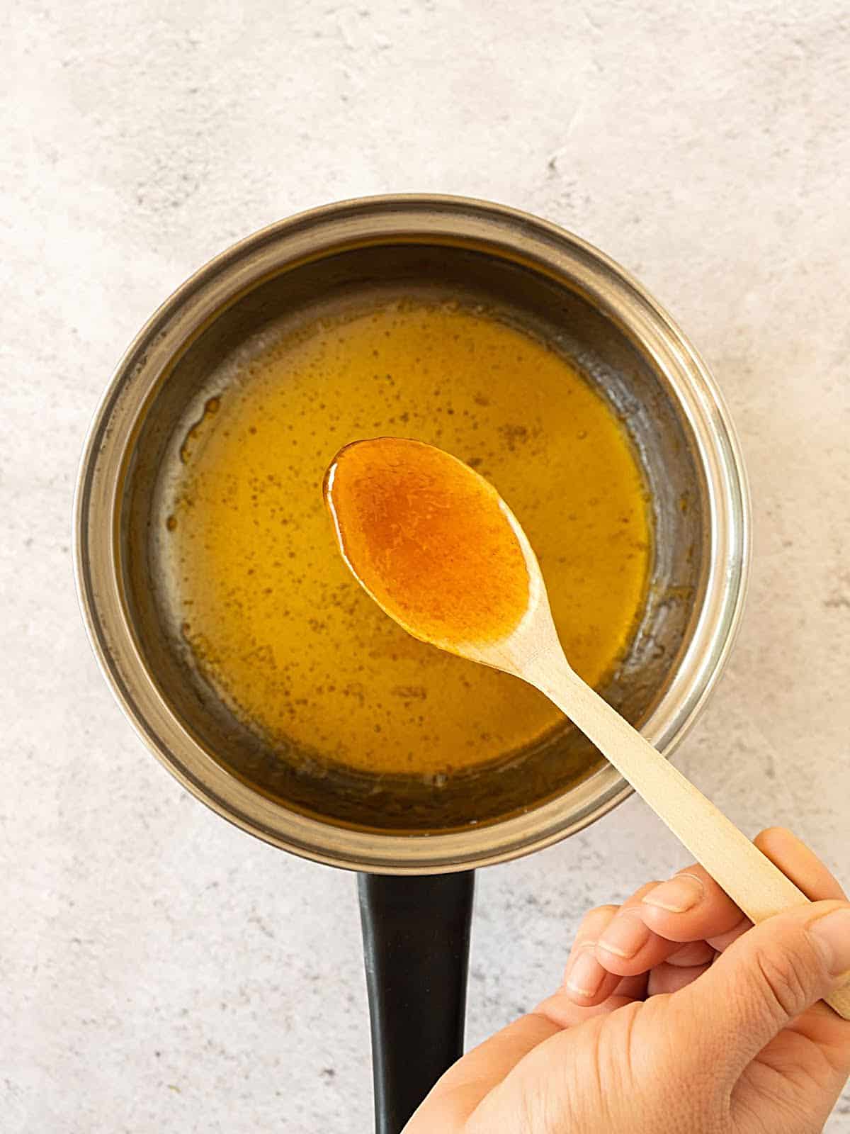 Wooden spoon lifting honey sriracha glaze from a saucepan. Light grey surface.