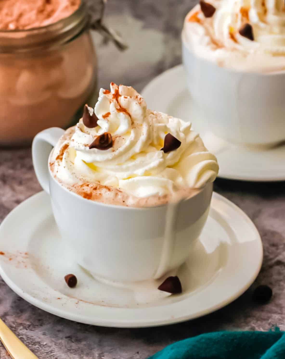 Hot chocolate with whipped cream in a white cup and saucer. Hot cocoa jar and cup in the background.