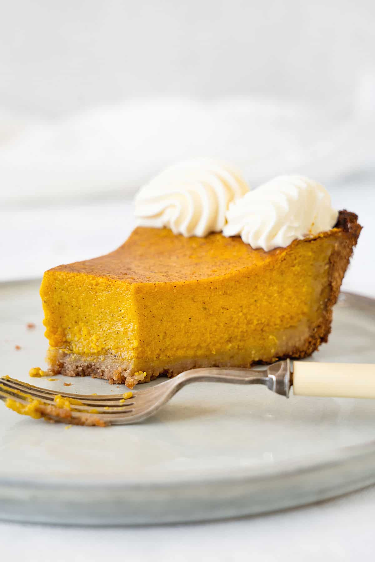 Slice of bitten pumpkin custard pie with whipped cream on a grey plate. A silver fork. Light grey background.