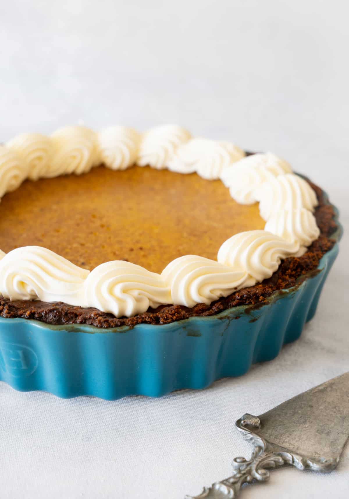 Partial view of teal pie dish with pumpkin cream pie. Light grey surface and background. Silver cake server. 