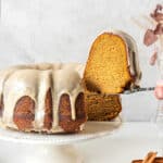 Glazed sweet potato bundt cake on a white cake plate. Slice being lifted. Light grey background.