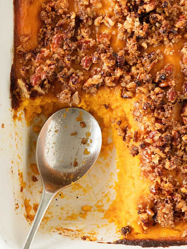 Close up of sweet potato pecan casserole in a white dish with a silver spoon. One missing serving.