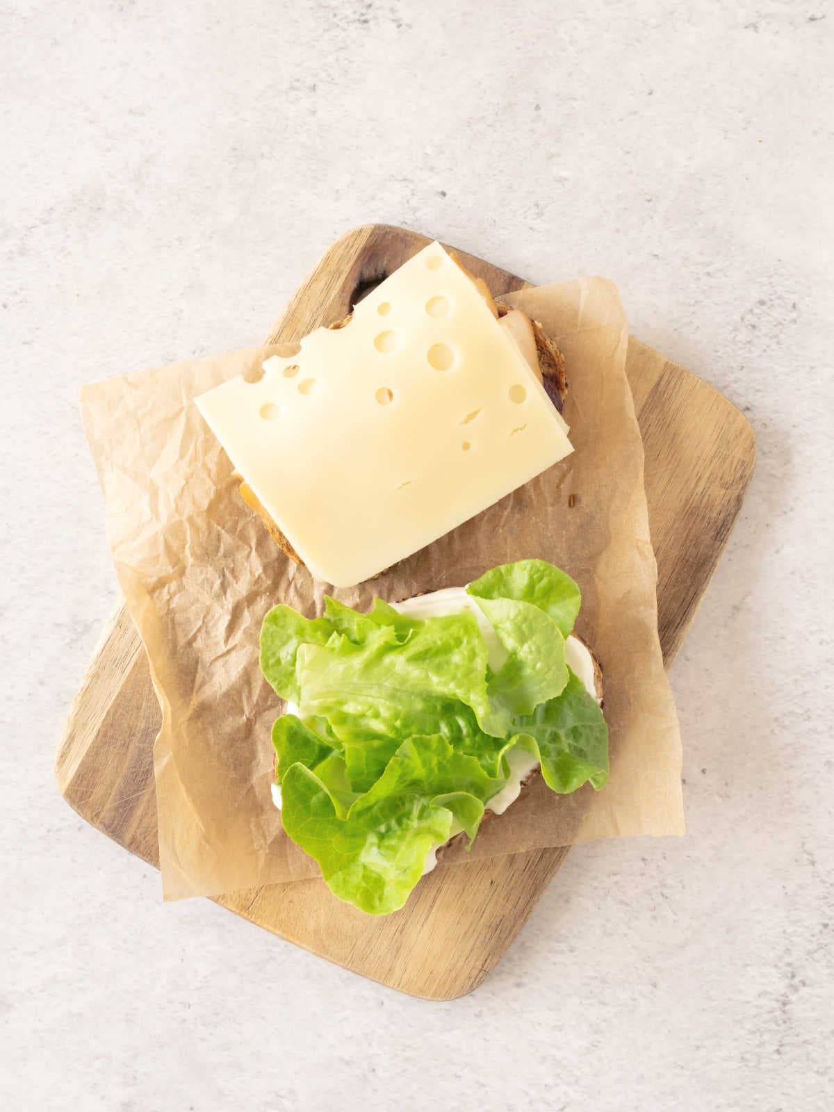 Grey surface with wooden board and sandwich being assembled with cheese and lettuce.