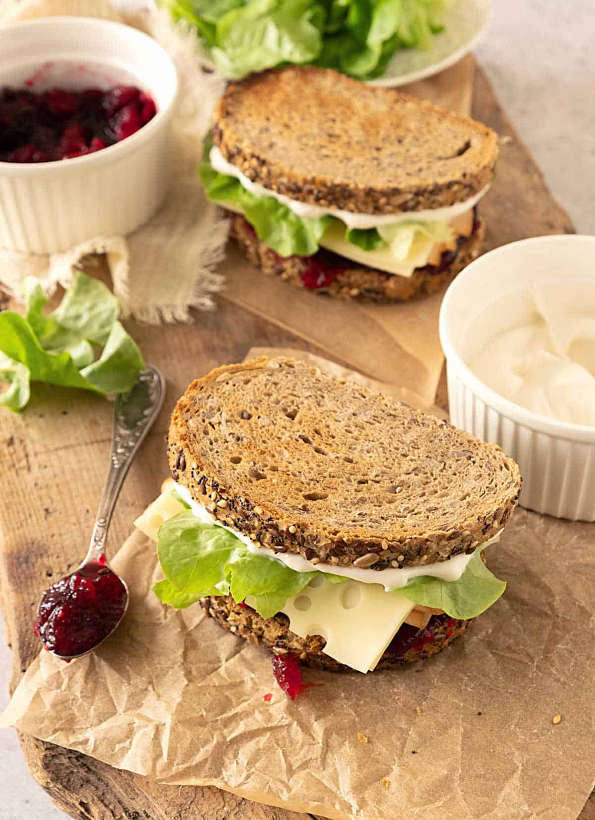 Two turkey lettuce cranberry sandwiches on beige paper. White bowl with mayo. 