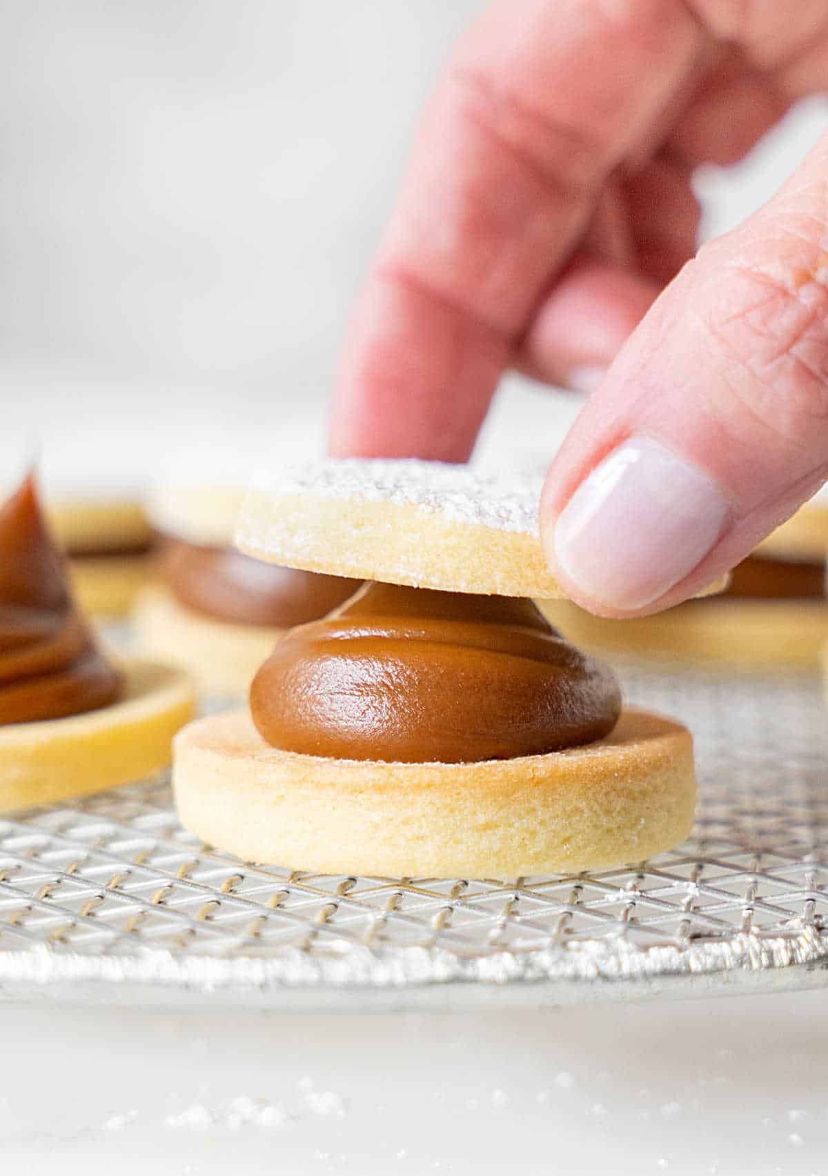 Placing top shortbread cookie over dulce de leche filled cookie on a wire rack. Light grey background.