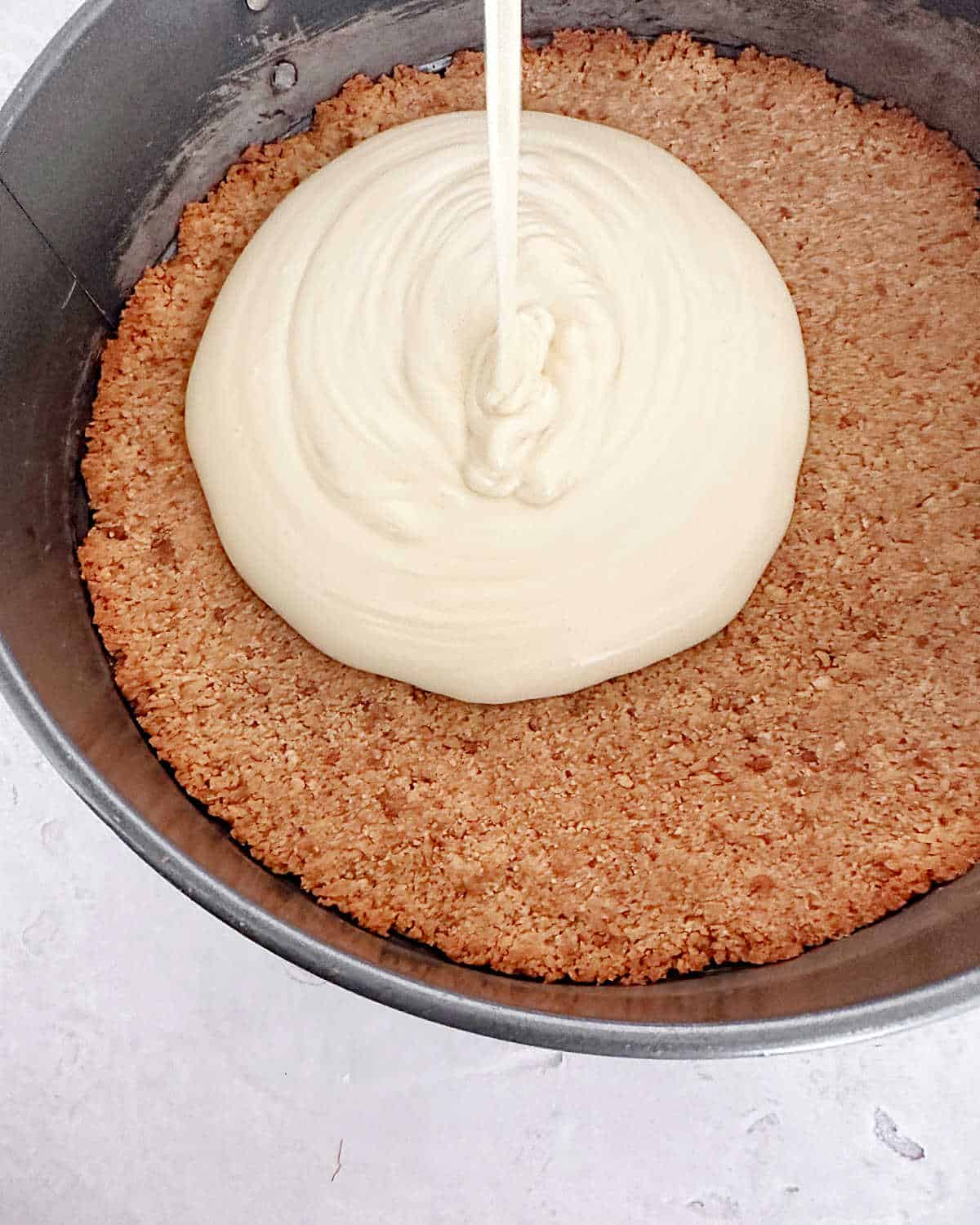 Cheesecake batter being poured on a graham crust in a metal pan. Light grey surface.