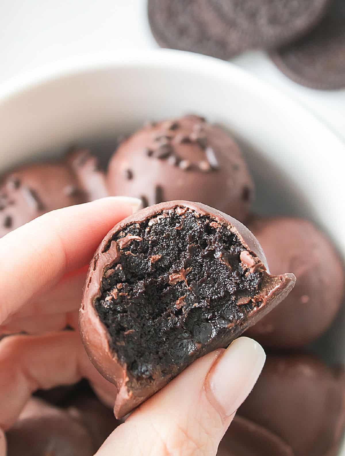 Hand holding a bitten Oreo truffle over a white bowl with more truffles.