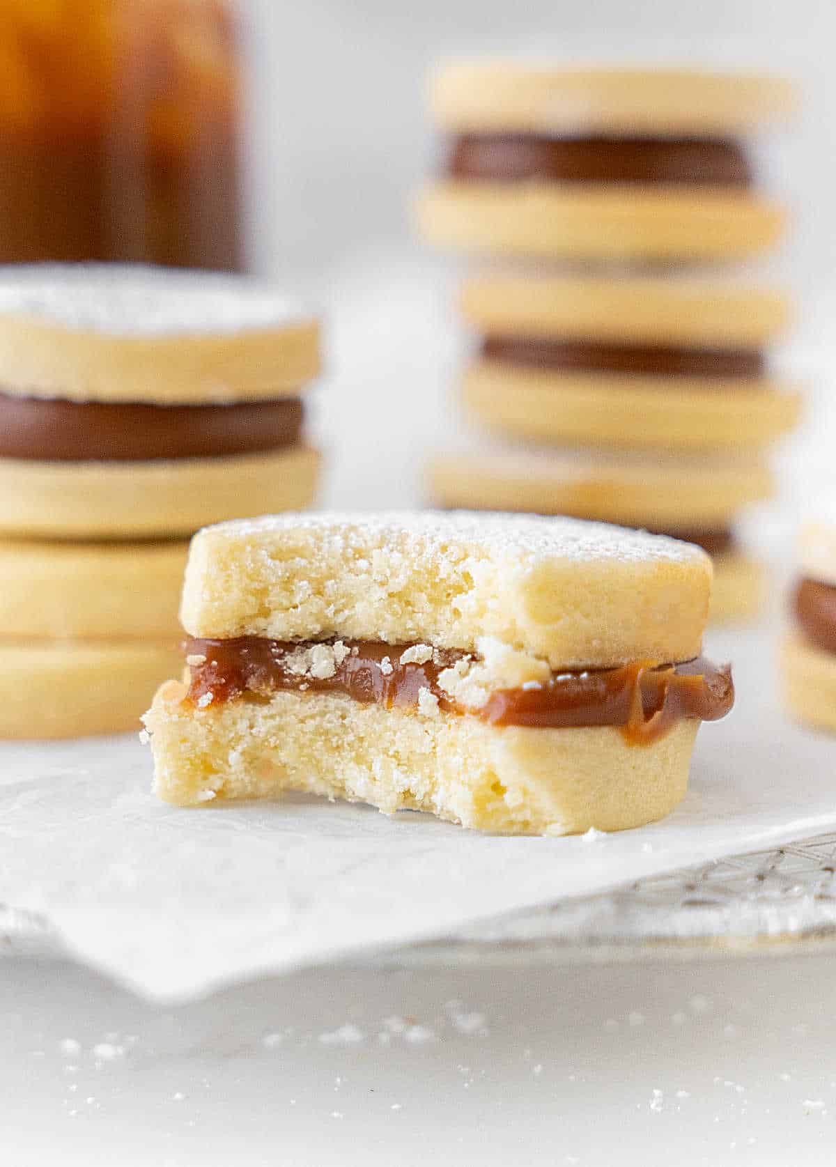 Bitten shortbread dulce de leche cookie on a white surface. Stack of alfajores in the background. 