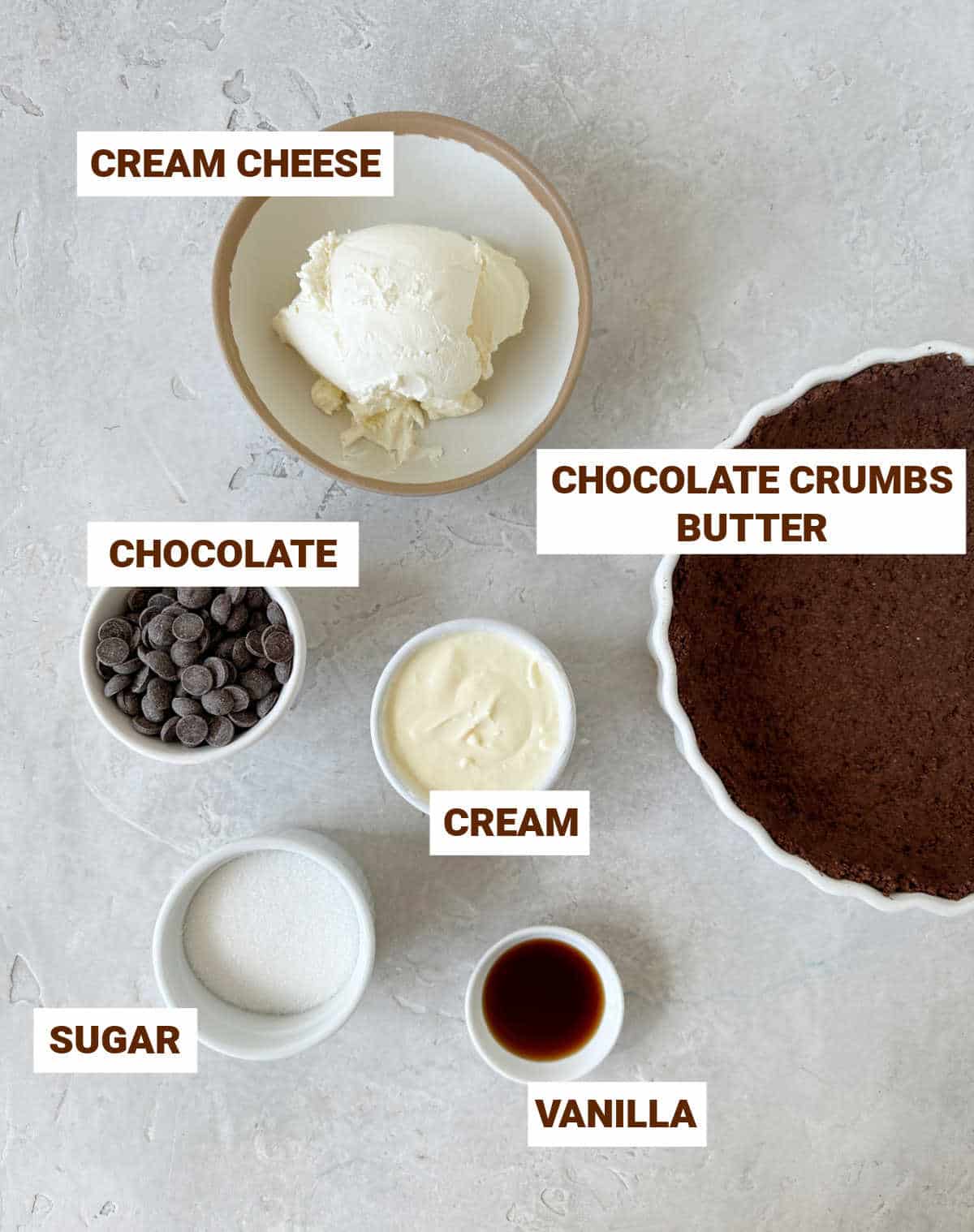 Light grey surface with bowls containing ingredients for chocolate cream pie including chocolate crust, cream cheese, vanilla and sugar.