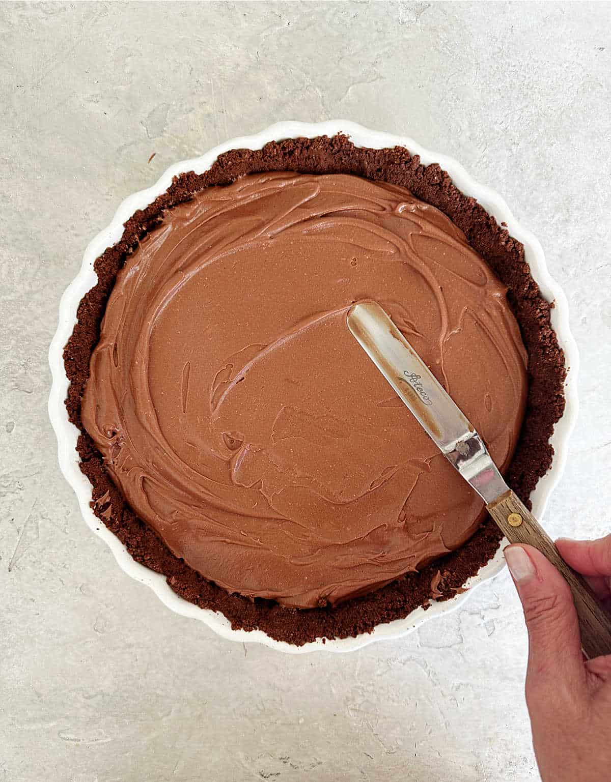 Spreading chocolate cheesecake filling on a chocolate crust with an offset spatula. White dish on grey surface.