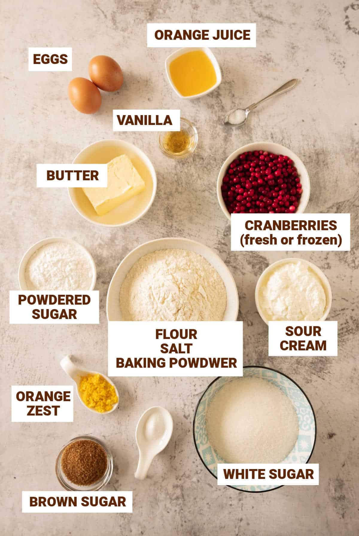 Gray surface with bowls containing ingredients for cranberry cake including butter, sour cream, sugars, eggs, orange, flour mixture.