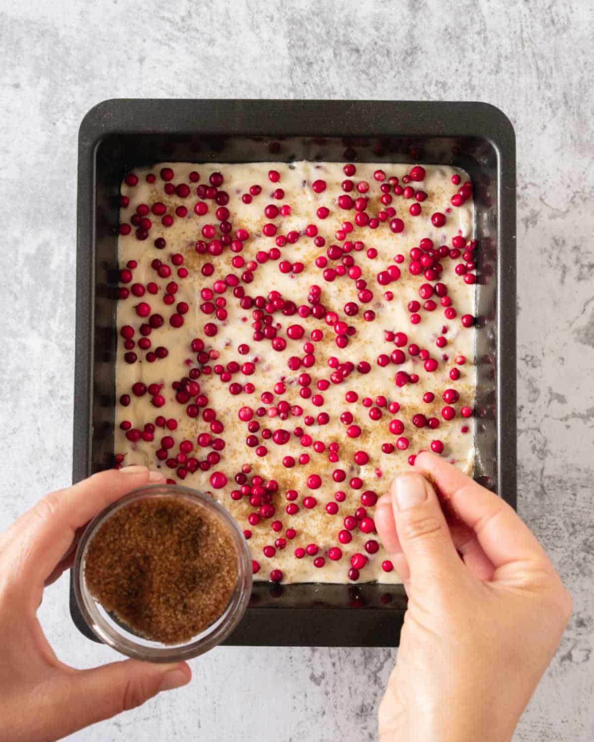 Sprinkling brown sugar on top of fresh cranberries on coffee cake batter in a metal pan. Gray surface.