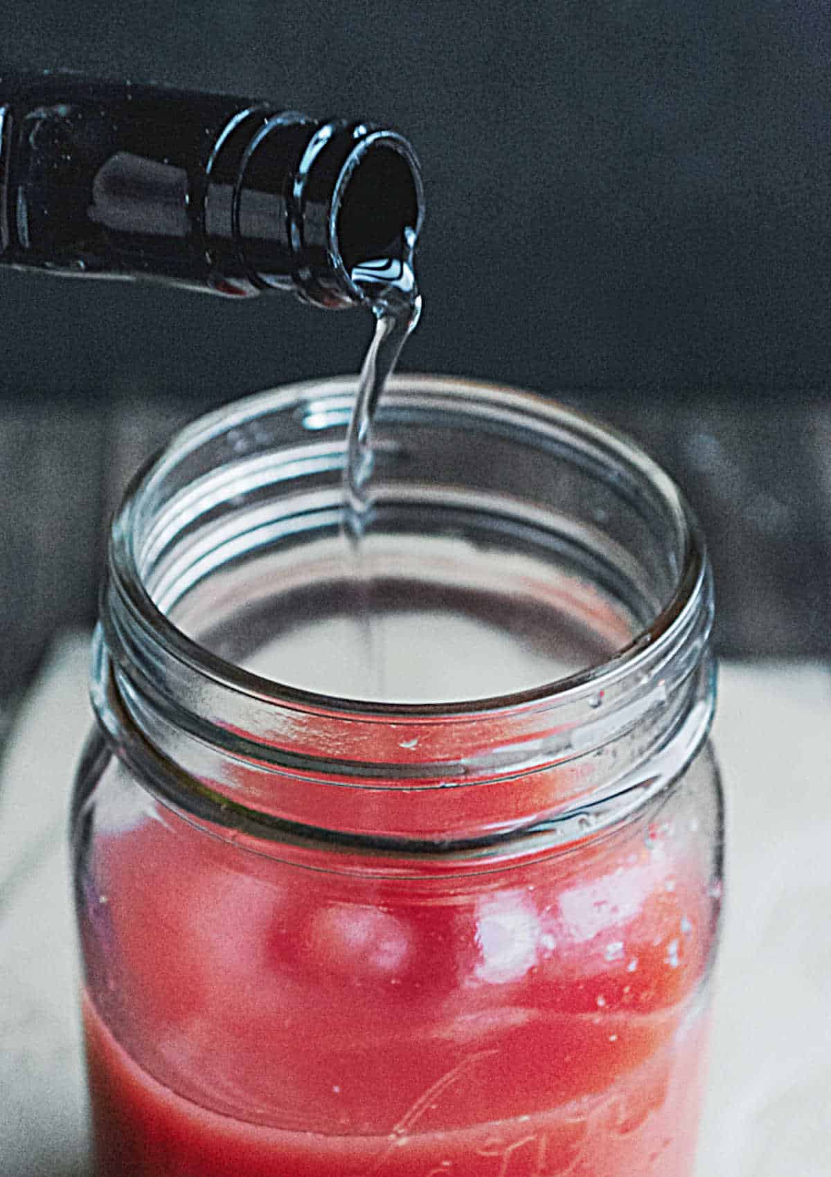 Glass mason jar with cranberry juice. Vodka being poured into it. White surface and black background.