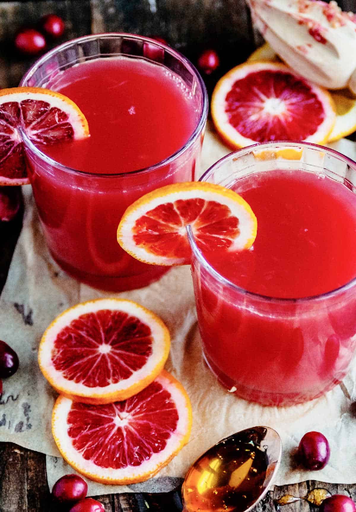 Orange slices and cranberries around two glasses with cranberry orange cocktail on a white and wood surface.