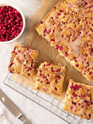 View from above of fresh cranberry coffee cake on a beige paper and wire rack. Bowl of cranberries. Light grey surface.