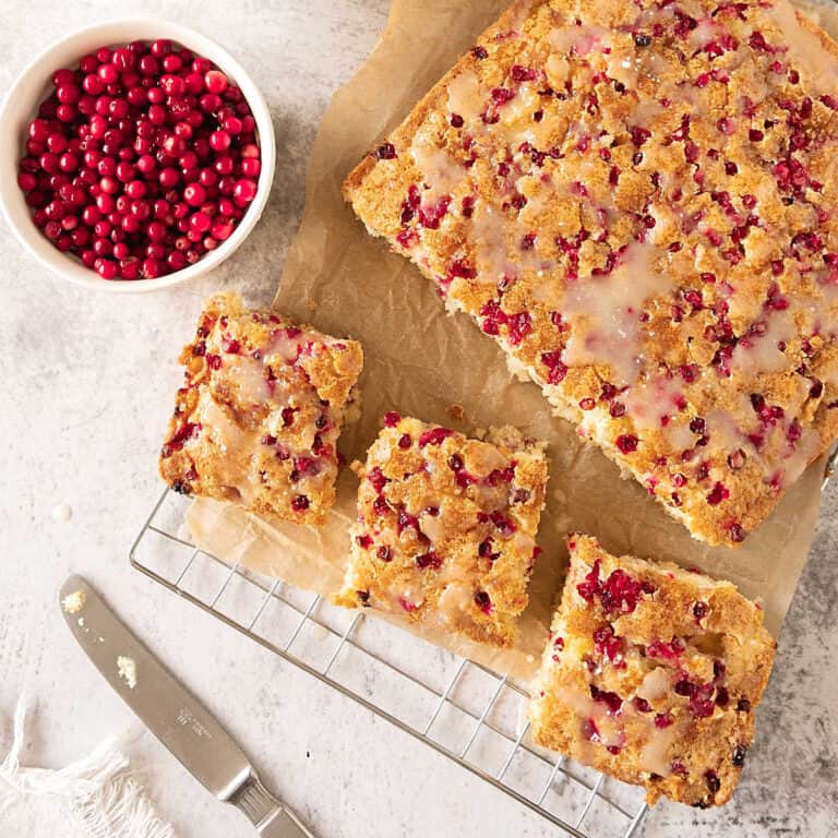 View from above of fresh cranberry coffee cake on a beige paper and wire rack. Bowl of cranberries. Light grey surface.