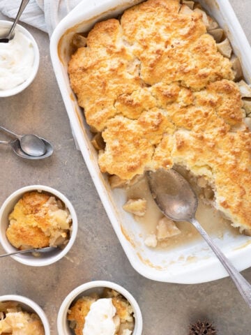 Apple cobbler in a white rectangular dish. Servings in white bowls. Brownish surface, spoons. Top view.