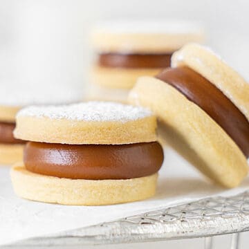 Two leaning shortbread dulce de leche cookies on a white paper and wire rack. Light grey background.