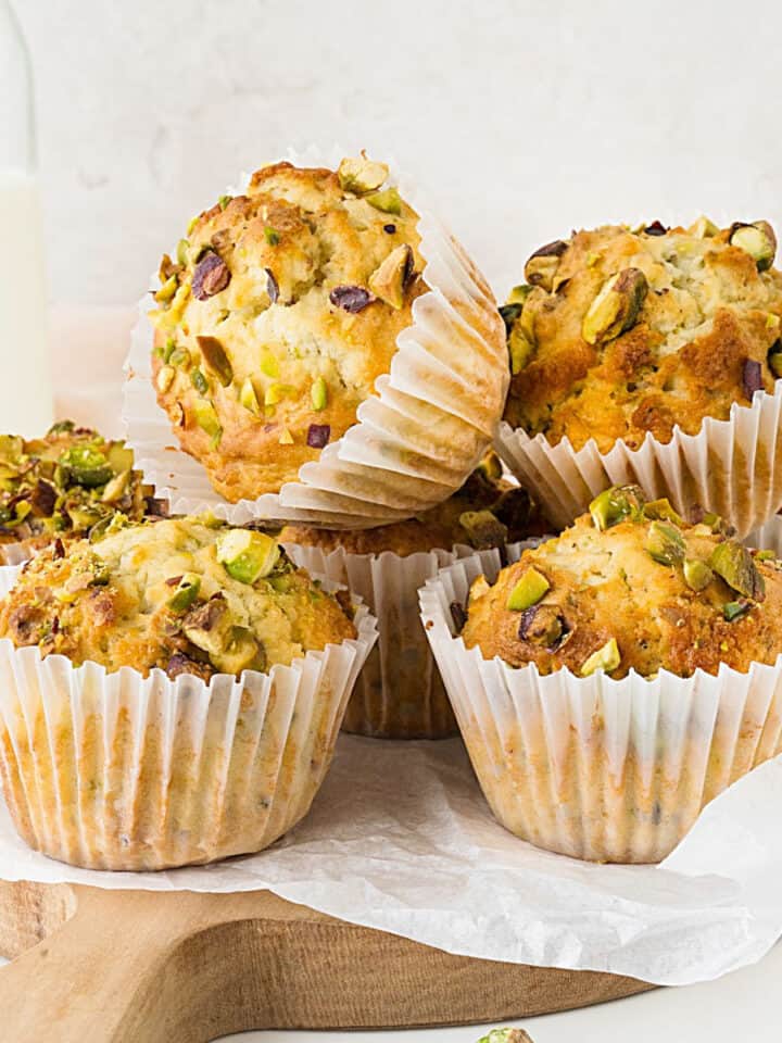 Pile of pistachio muffins in white paper liners on a wooden board with a white paper. Light grey background.