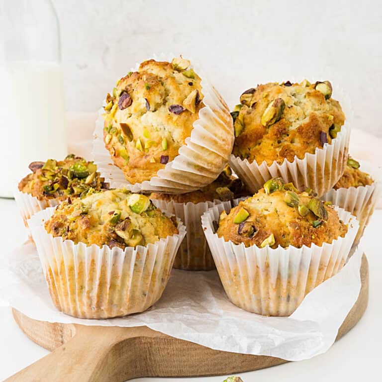 Pile of pistachio muffins in white paper liners on a wooden board with a white paper. Light grey background.