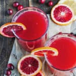Two glasses with cranberry cocktail and orange slices. Wood and white paper surface.