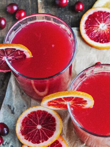 Two glasses with cranberry cocktail and orange slices. Wood and white paper surface.