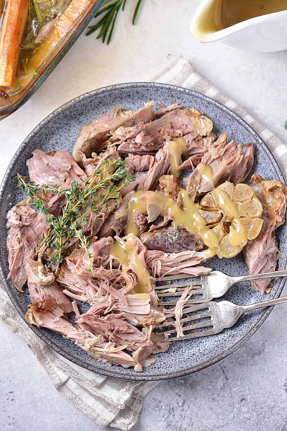 Serving of pulled-apart leg of lamb with gravy on a grey plate. Silver forks, light grey surface, striped cloth, gravy boat. 