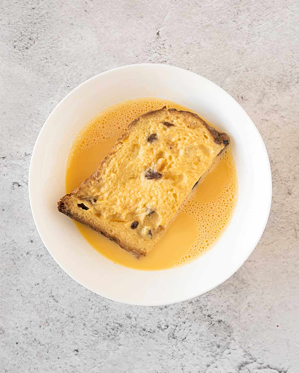 Top view of white bowl with egg mixture and slice of panettone. Gray surface.