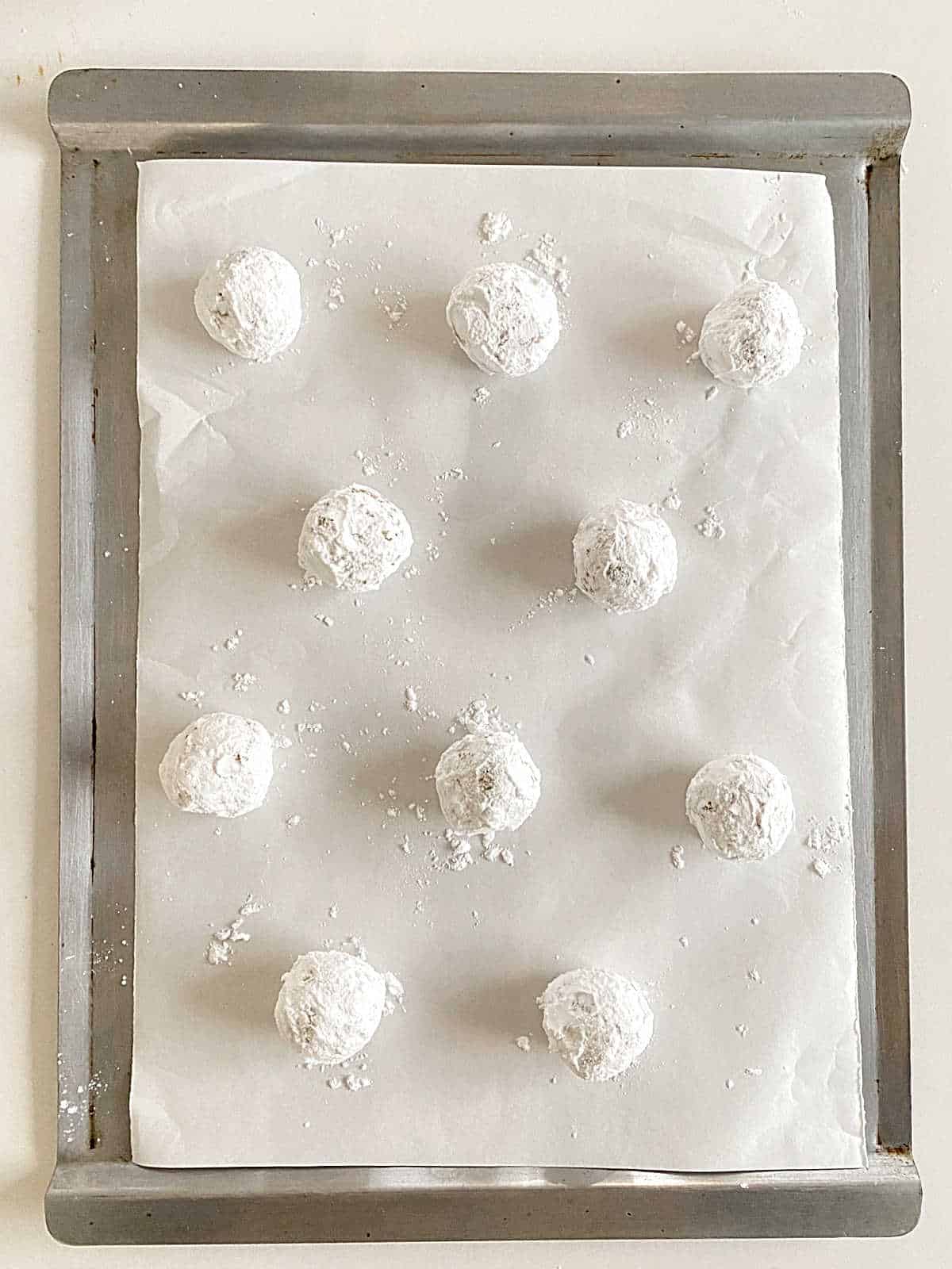 Metal pan with white parchment paper and powdered sugar crinkle cookies before baking.