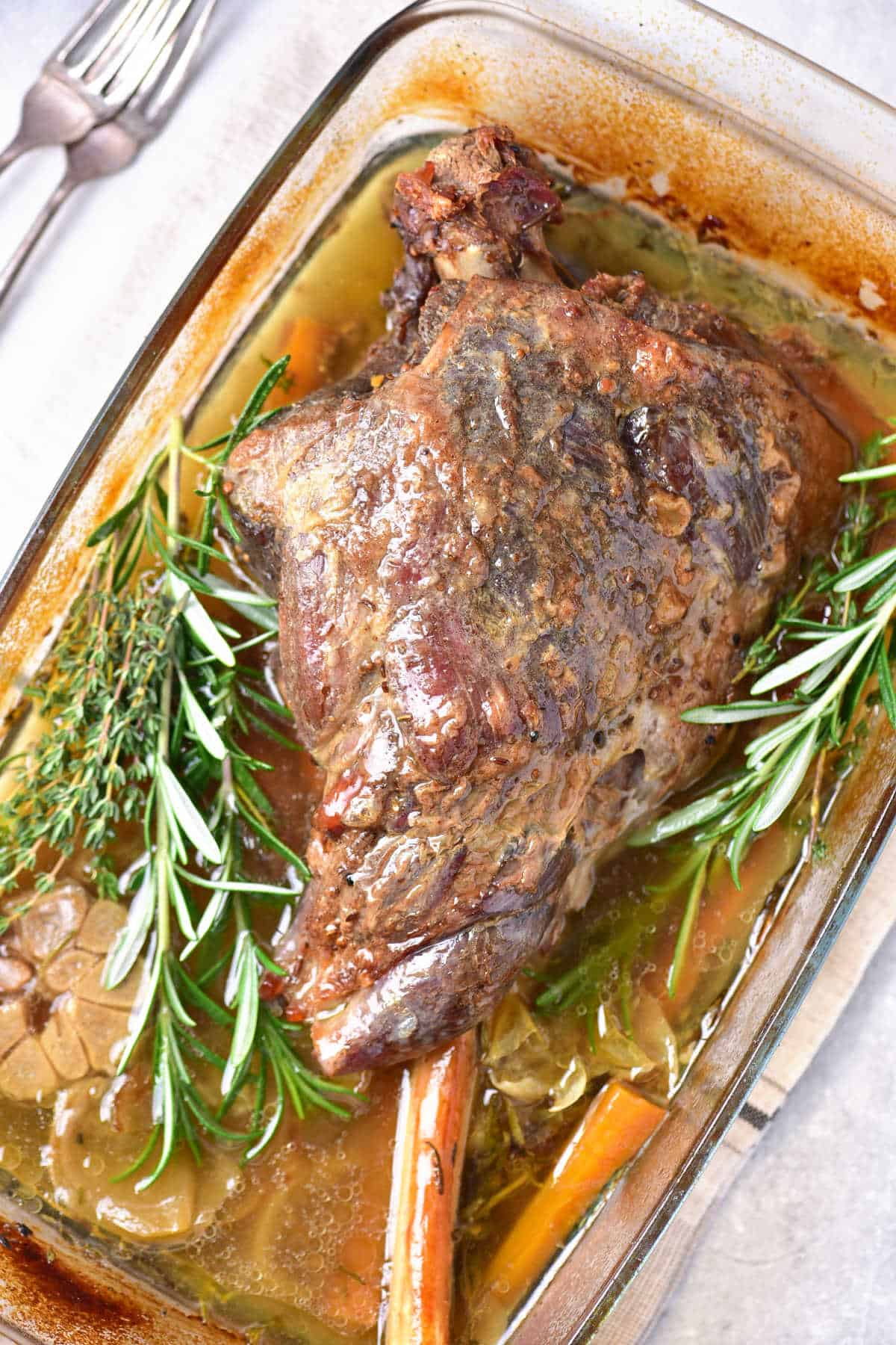Fresh herbs around a juicy roasted leg of lamb in a glass dish.