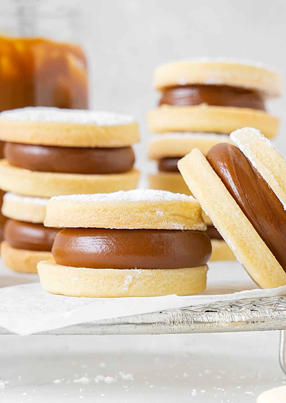 Dulce de leche shortbread alfajores stacked on white paper and wire rack. Light gray background. 
