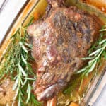 Whole slow roasted leg of lamb with fresh herbs on a glass dish.