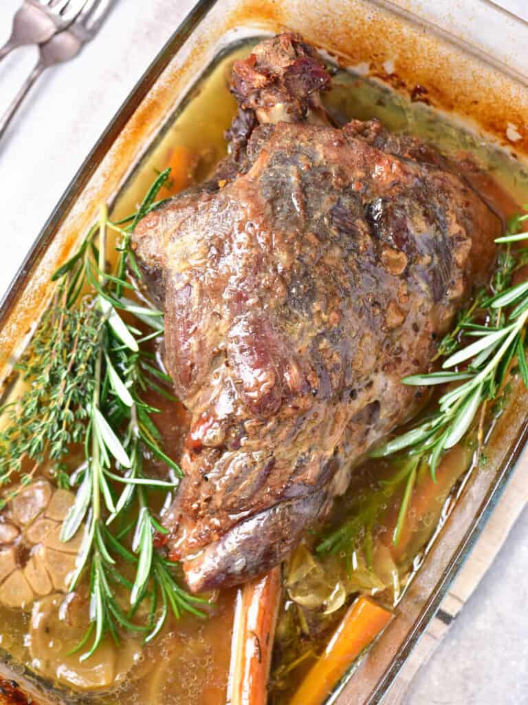 Whole slow roasted leg of lamb with fresh herbs on a glass dish.