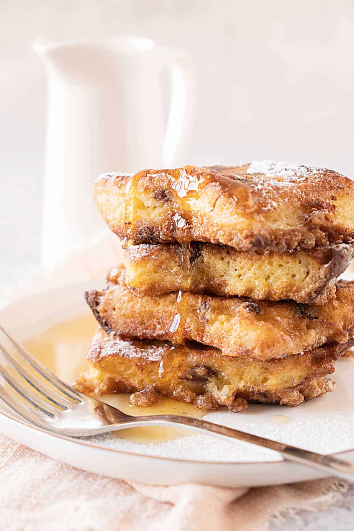 White plate with stack of panettone French toast with syrup. Silver fork, white jug in the white background.