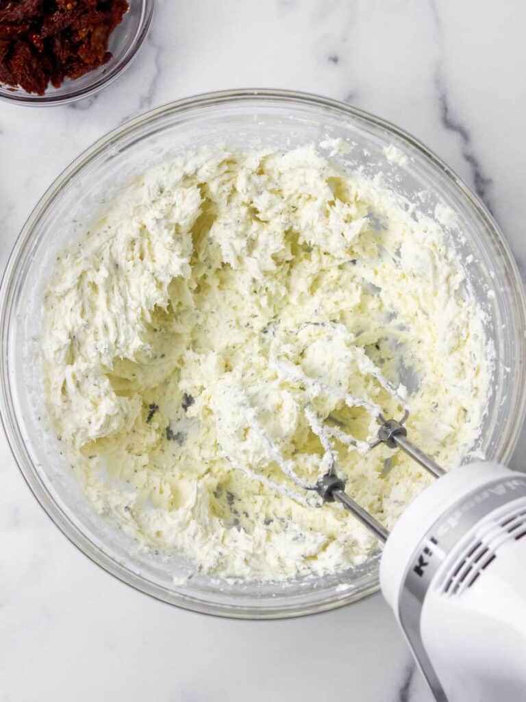 Glass bowl with cheese herb mixture being beaten. White marbled surface.