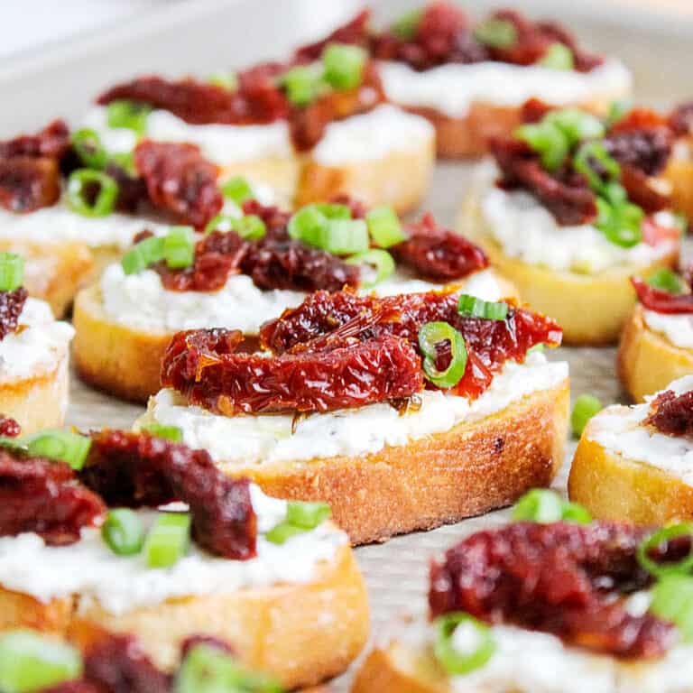 Close up bruschettas with cheese spread, dried tomatoes and chopped green onions on a metal pan.