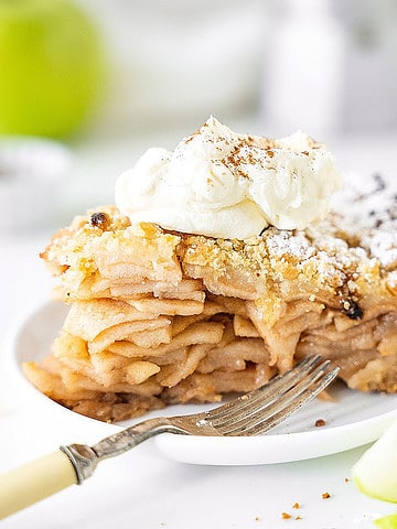 Cream-topped slice of apple crumb pie on a white plate. A silver fork, green apple pieces. White background.