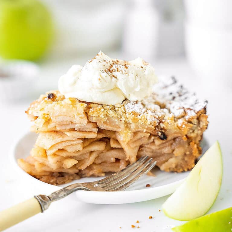 Cream-topped slice of apple crumb pie on a white plate. A silver fork, green apple pieces. White background.