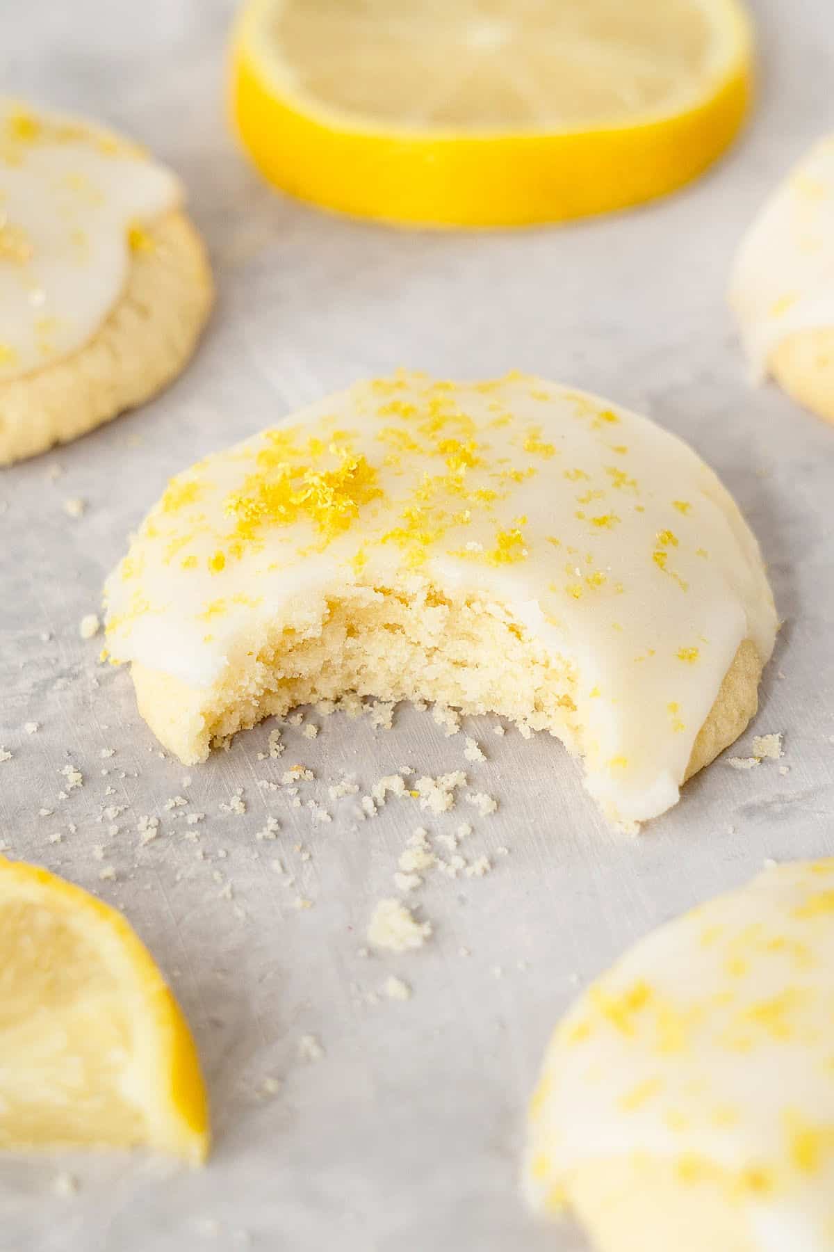 Bitten lemon ricotta cookie with glaze. Gray surface, lemon slices, more cookies.