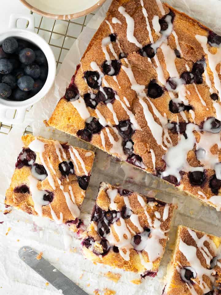 Glazed blueberry coffee cake, whole and in squares on a wire rack. White surface, bowl of blueberries. Top view.
