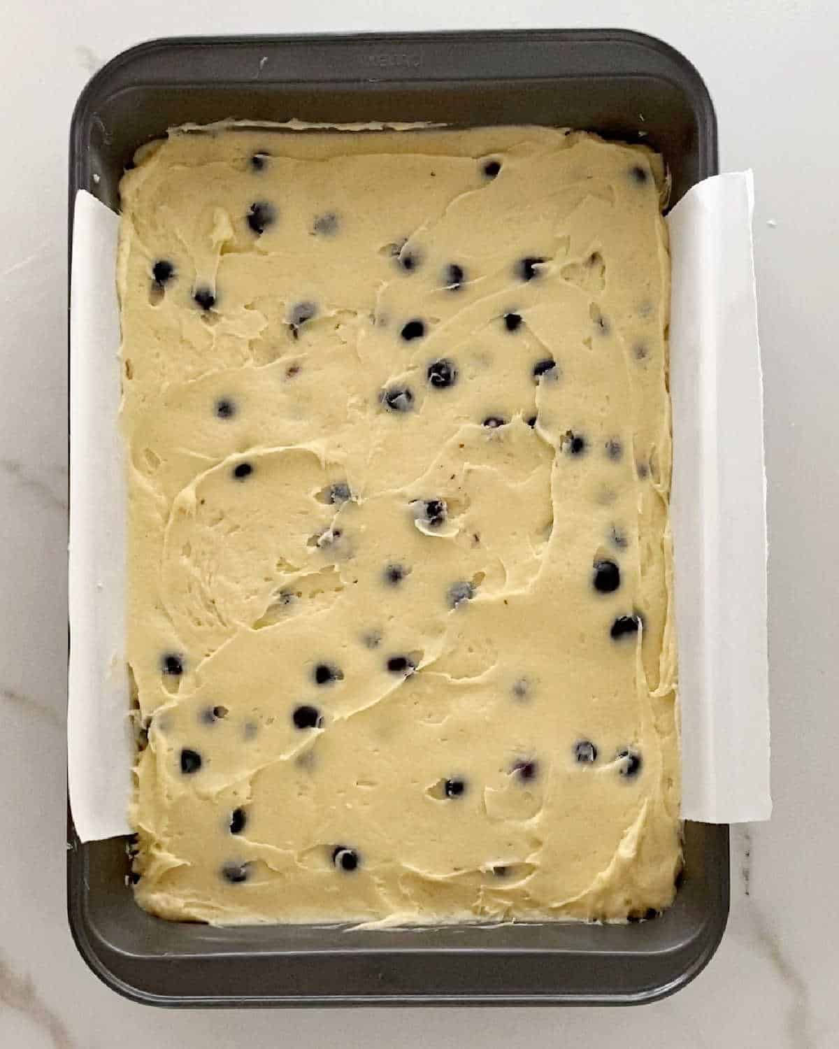 Batter for blueberry coffee cake in a metal rectangular pan. White surface.