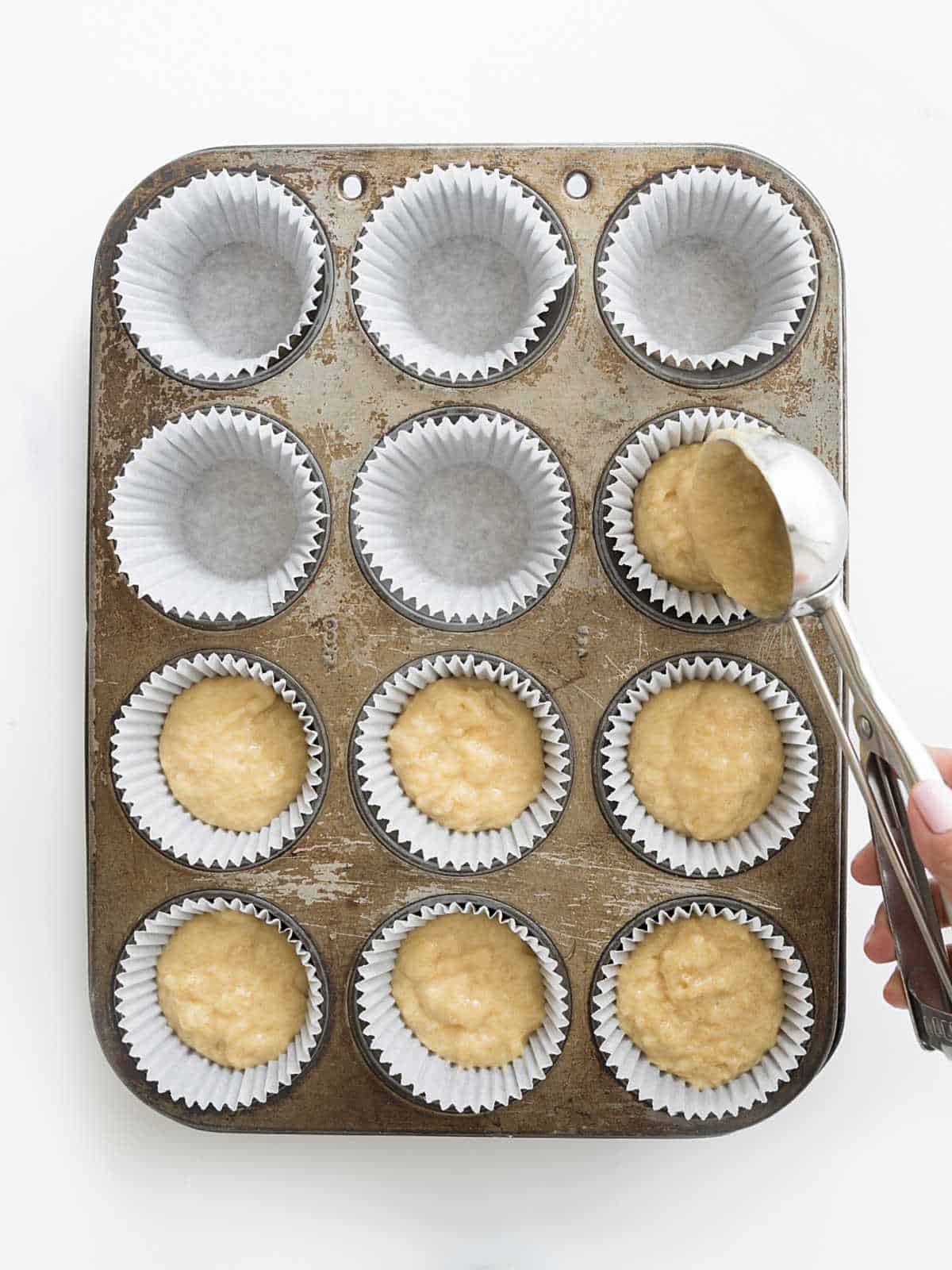 Scooping muffin batter into white paper liners on a metal muffin pan. White surface.
