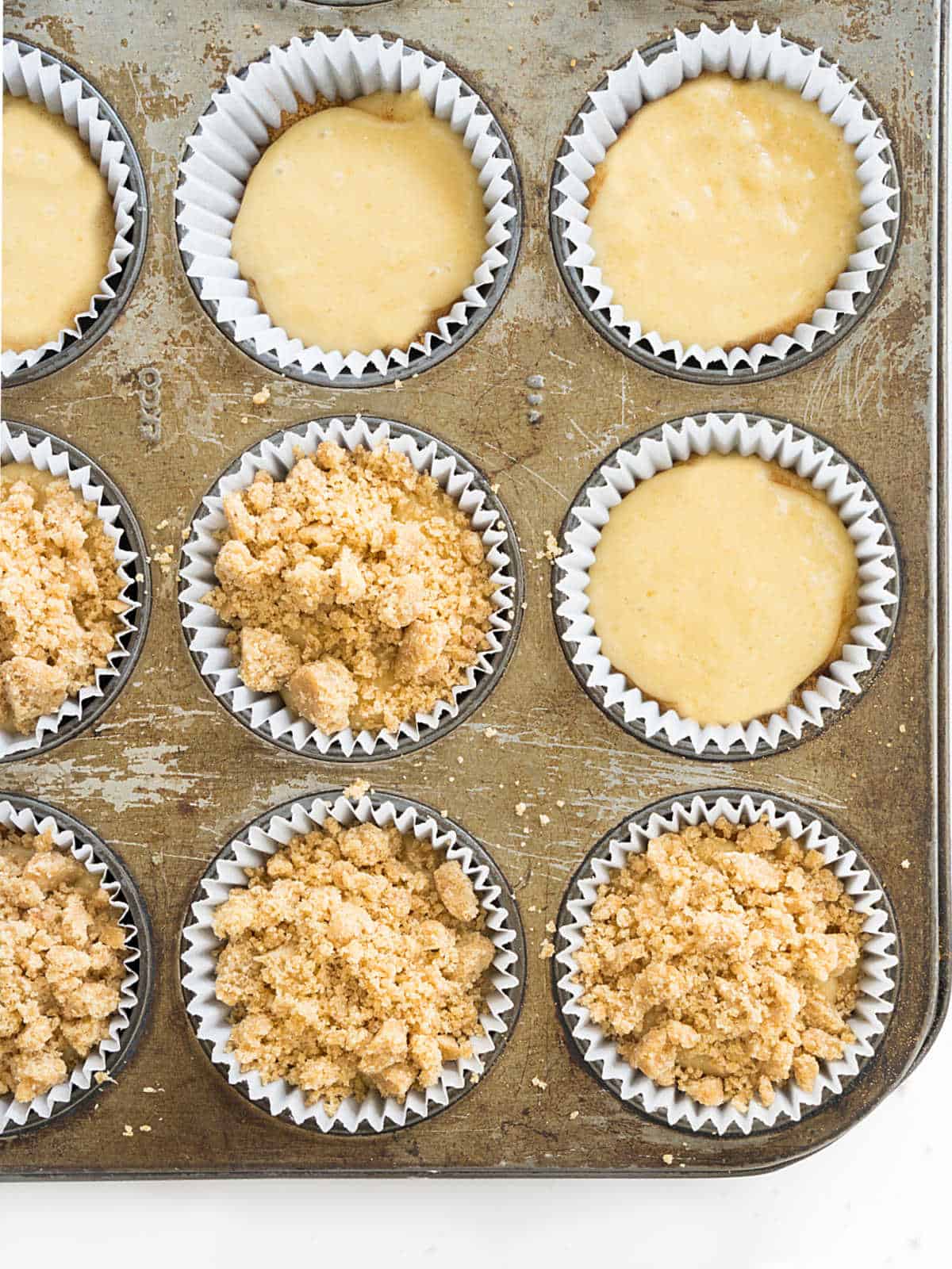 Metal muffin pan with unbaked cinnamon muffins, some with crumble, some plain. 