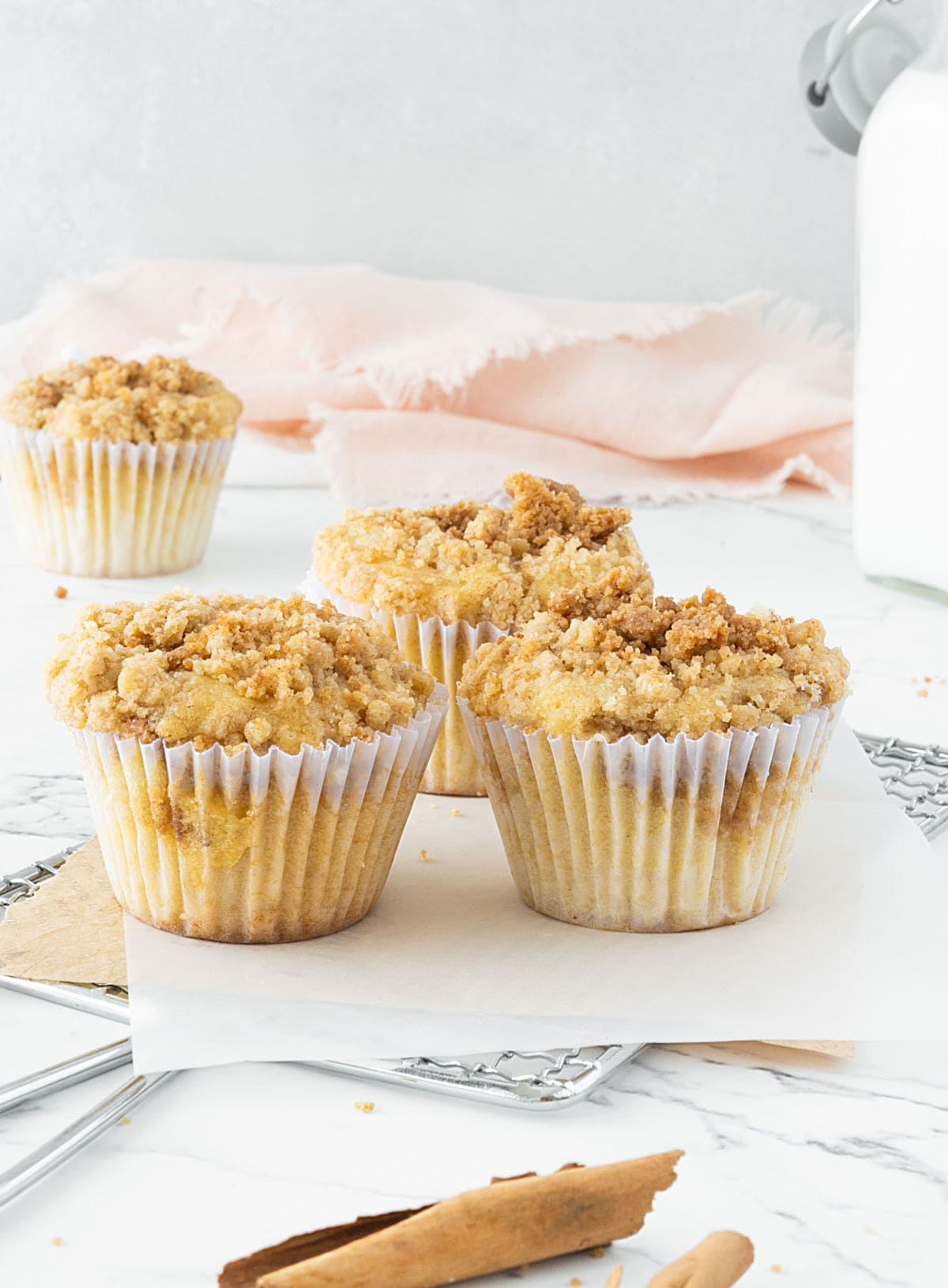 Several crumb topped cinnamon muffins on paper liners and white paper. Pink cloth, grey background, white marbled surface.