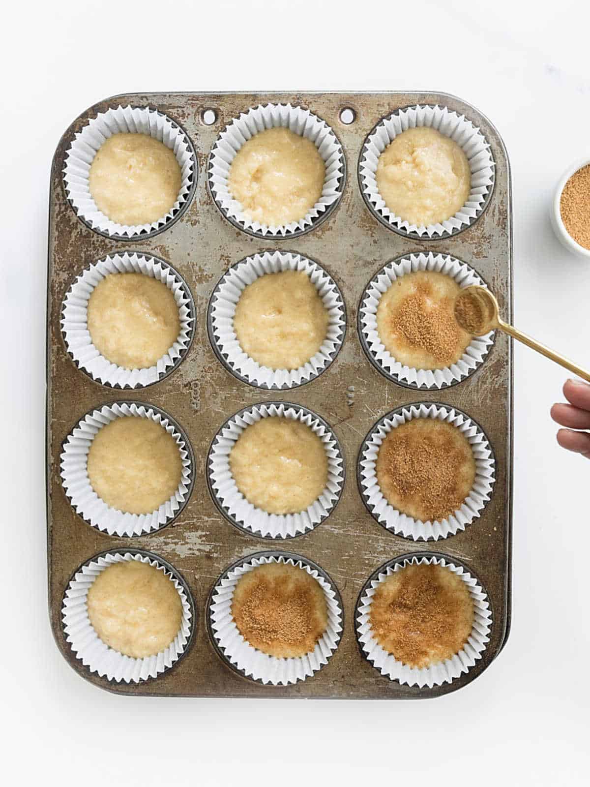 Adding cinnamon sugar to muffin batter in paper liners on a metal pan.