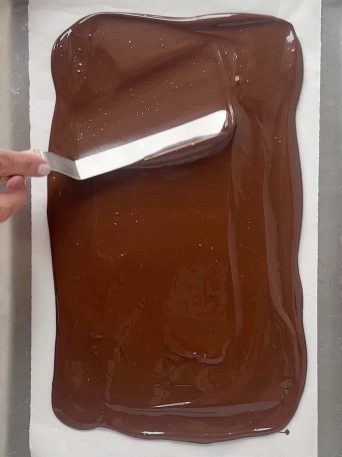 Spreading melted dark chocolate with an offset spatula on white parchment paper.