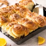 Close up dark oven tray of glazed orange cinnamon rolls on a gray surface. Orange slices.