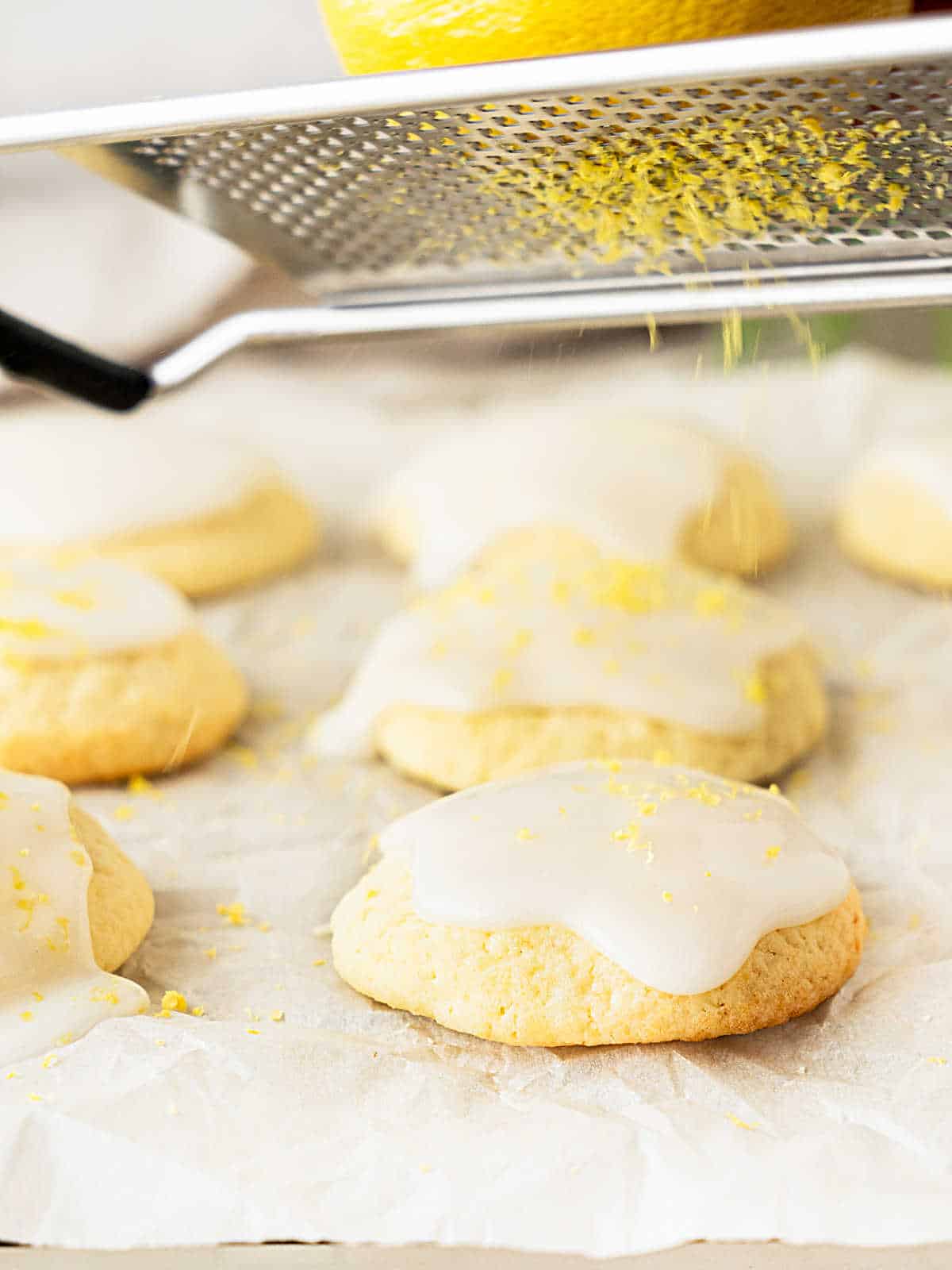 Zesting lemon over glazed ricotta cookies on white parchment paper.