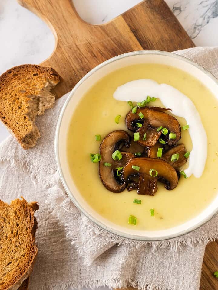 Potato, leek and mushroom soup in a white bowl on a beige linen. Wooden board, white marble, toast pieces.