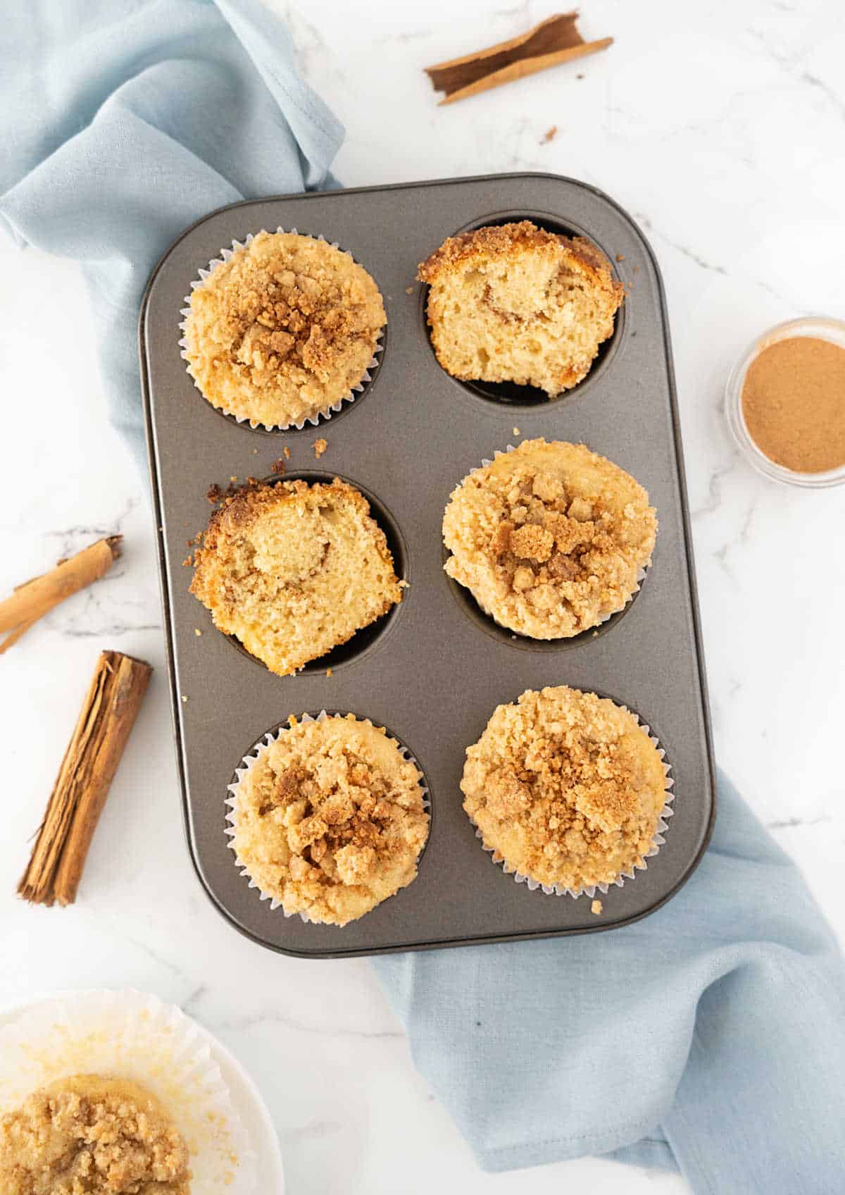 Top view of metal pan with halved and whole cinnamon crumble muffins. White surface, light blue linen. 